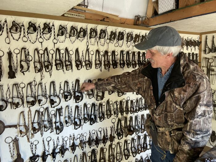An older man with white hair and a ball cap points to dozens of traps hung on a wall.