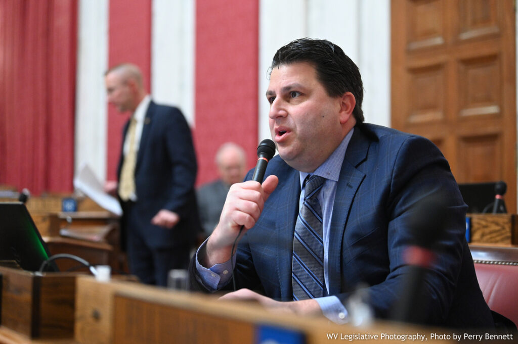 A man in a blue suit and tie speaks into a microphone from a desk in the West Virginia House of Delegates chamber.