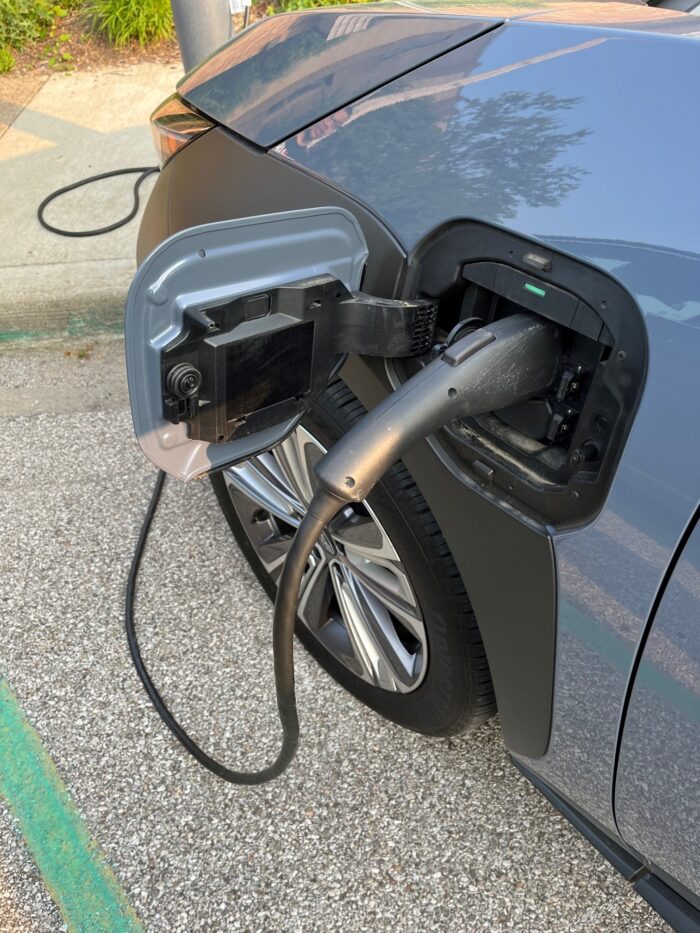 A black EV charger cord is plugged into a gray Subaru Solterra in the parking lot of a public library on a summer evening.