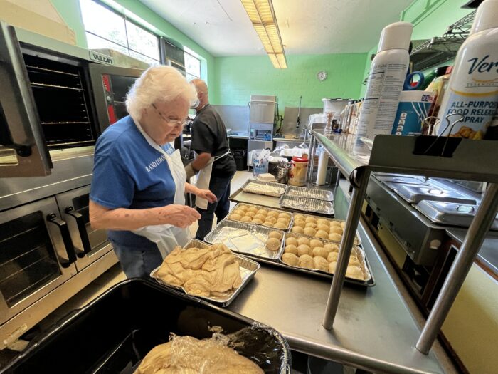 An elderly woman with white hair, wearing an apron, works with soft dough for pepperoni rolls. She wears glasses and a blue t-shirt.
