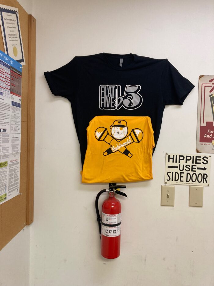A black and yellow shirt hang on a wall. Next to them is a sign that reads, "Hippies Use Side Door."