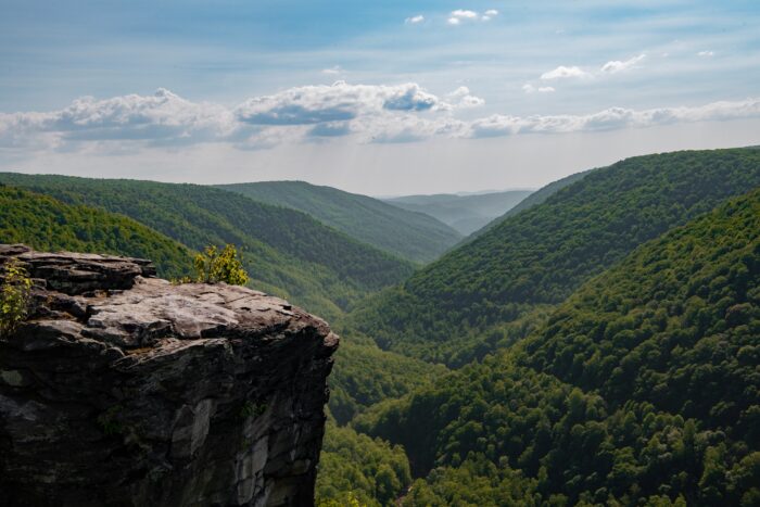 Sunlight streams through white clouds into a forested valley between rolling hills. In the foreground a rocky outcropping can be seen.