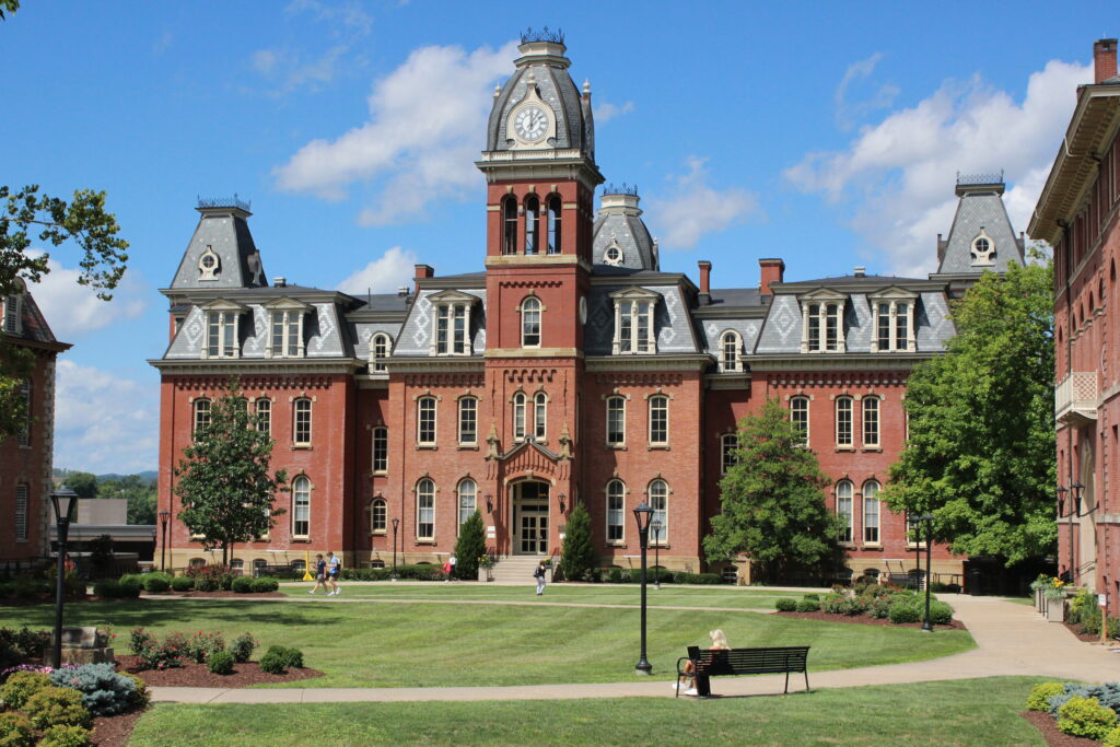 West Virginia University's iconic Woodburn Hall - a brick building with a distinctive clock tower - on a sunny day in front of a blue sky with several clouds. In front of the building can be seen the green space Woodburn Circle, with students walking across.