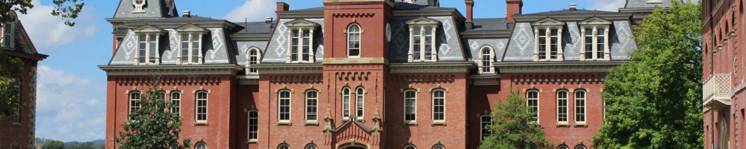 West Virginia University's iconic Woodburn Hall - a brick building with a distinctive clock tower - on a sunny day in front of a blue sky with several clouds. In front of the building can be seen the green space Woodburn Circle, with students walking across.