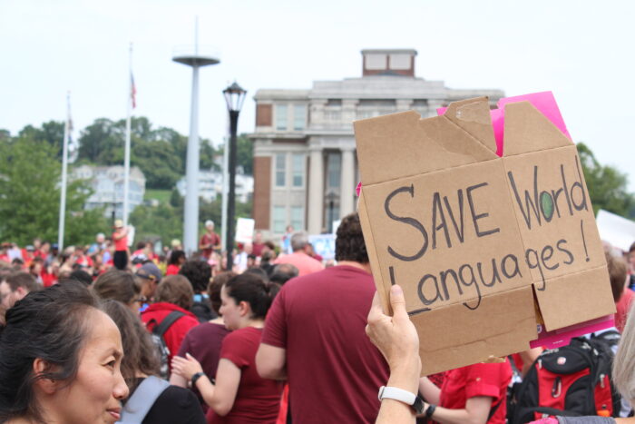 A cardboard sign reading "Save World Languages" is held up in a crowd of red shirts. In the background can be seen WVU's Oglebay Hall