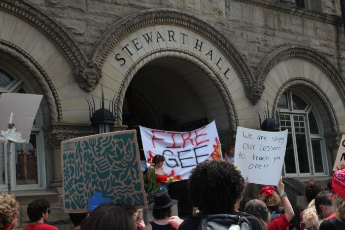 A sign on a bedsheet that reads "Fire Gee" over painted flames is held up under the entry arch of WVU's Stewart Hall. Another visible sign is held up over a crowd's head reads "We skipped our lessons to teach you one"