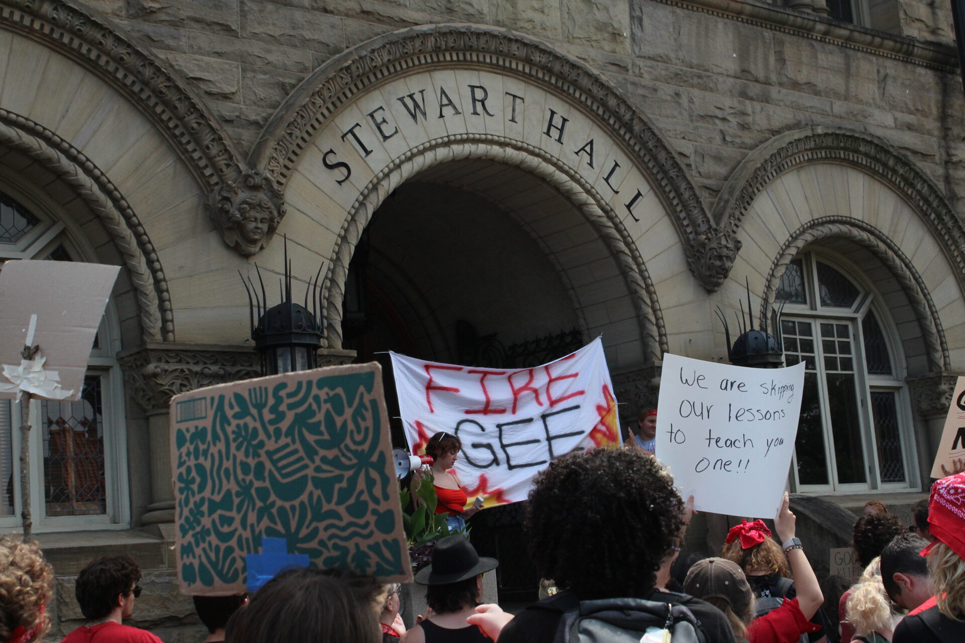 A sign on a bedsheet that reads "Fire Gee" over painted flames is held up under the entry arch of WVU's Stewart Hall. Another visible sign is held up over a crowd's head reads "We are skipping our lessons to teach you one!!"