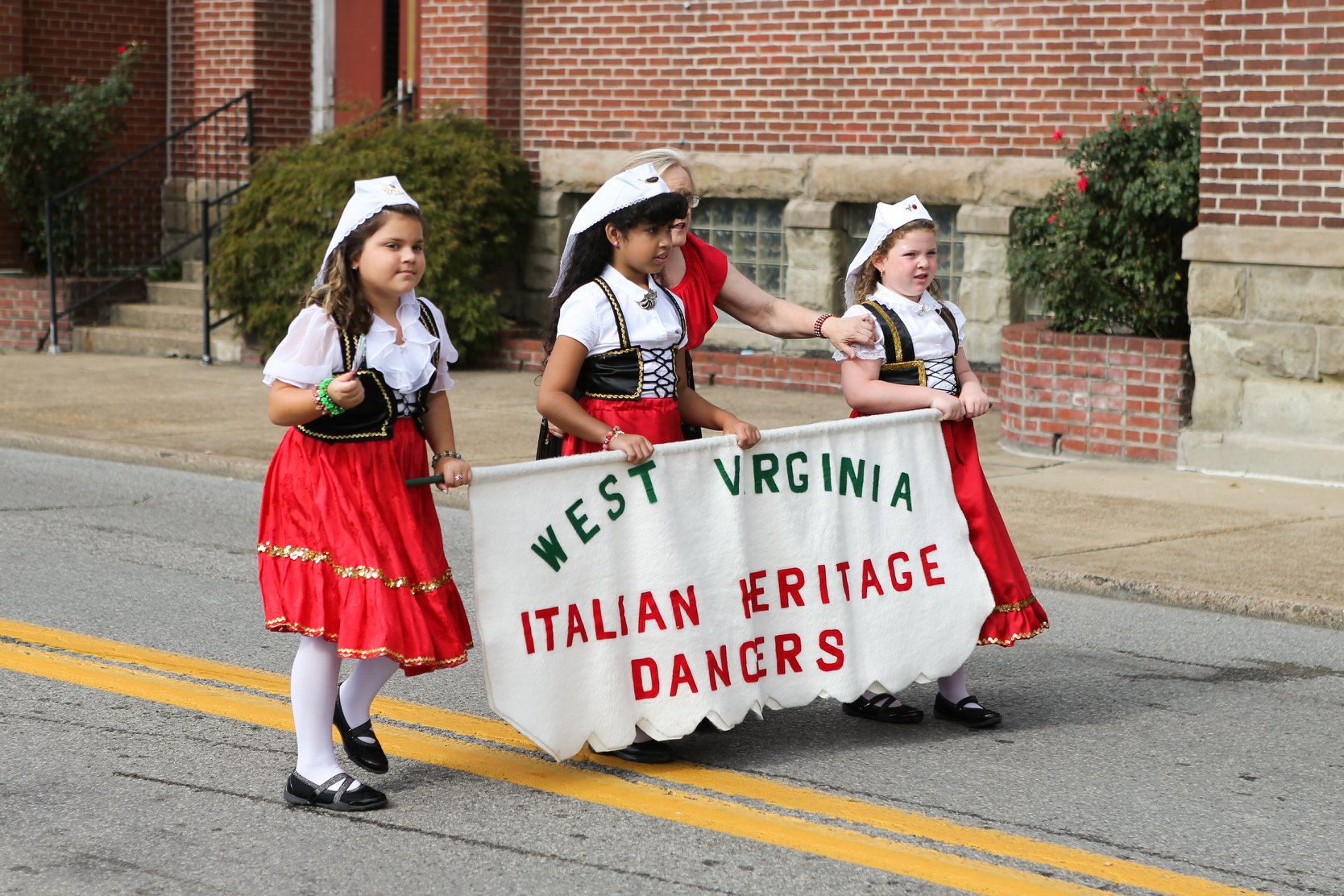 A group of children dressed in traditional Italian dress walk down a street holding a sign that reads West Virginia Italian Heritage Dancers.