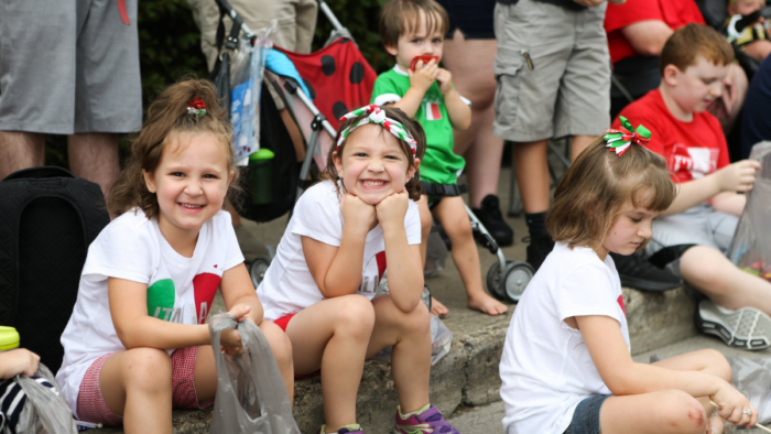 Two smiling girls wearing headbands in the Italian colors grin at the camera. They sit on the side of the street watching a parade.