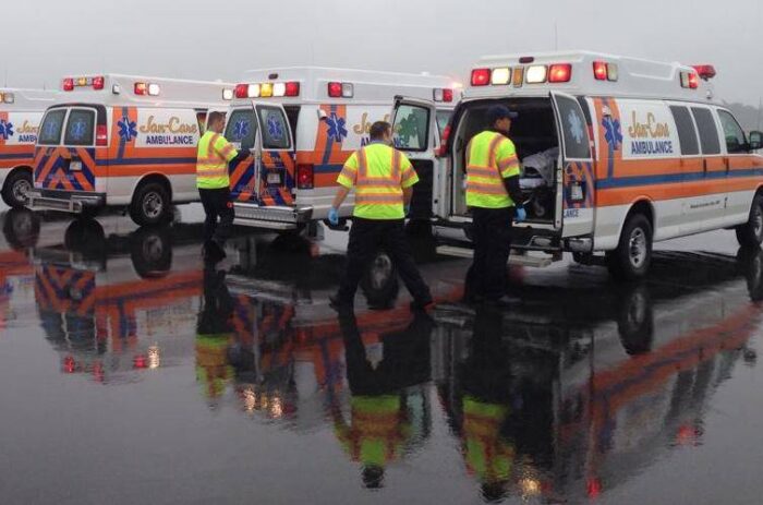 EMS workers load and unload three different ambulances parked in lots. The air is gray, and Drury and the pavement is wet. 