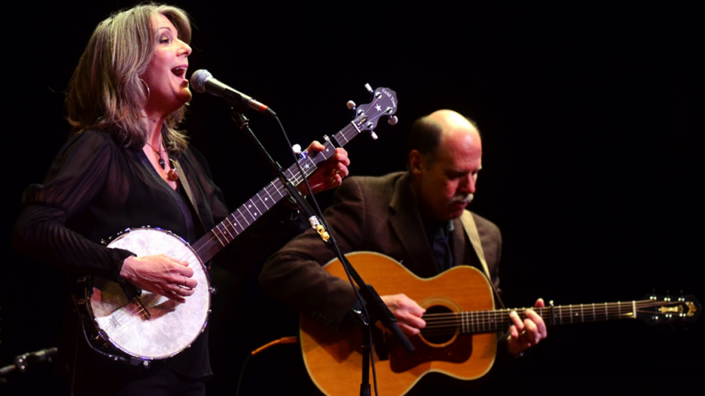 A woman with grey hair sings and plays the banjo while a man in the background plays the guitar.