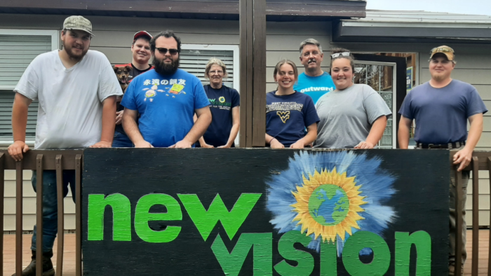 Eight people stand on a deck smiling for the camera. In front of them, along the railing, is a large sign that reads "New Vision" in bold green lettering. Painted just above it is the Earth surrounded by sun beams.