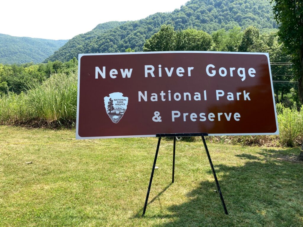 A brown highway sign with white lettering on a hazy August day with green mountain ridges in the background.