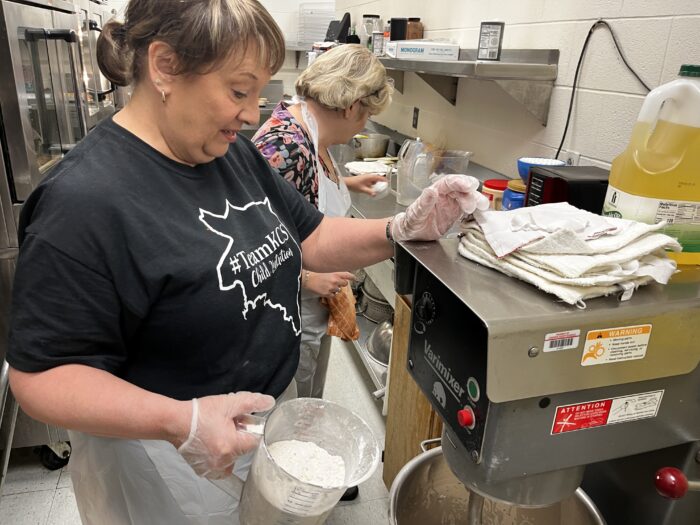 A woman in a black shirt and hairnet holds a pitcher full of flower. In the background is another woman in a hairnet and apron. They are in a school kitchen.
