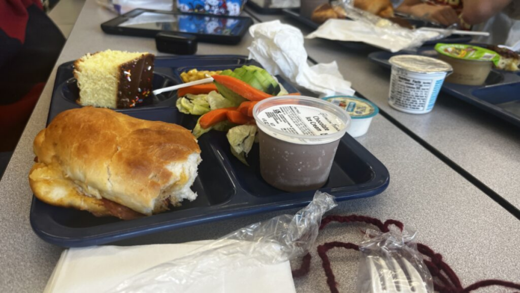 Close up of a fresh pepperoni roll is seen on a school tray. Shown also are vegetables, an ice cream cup, napkins, and a plastic fork.