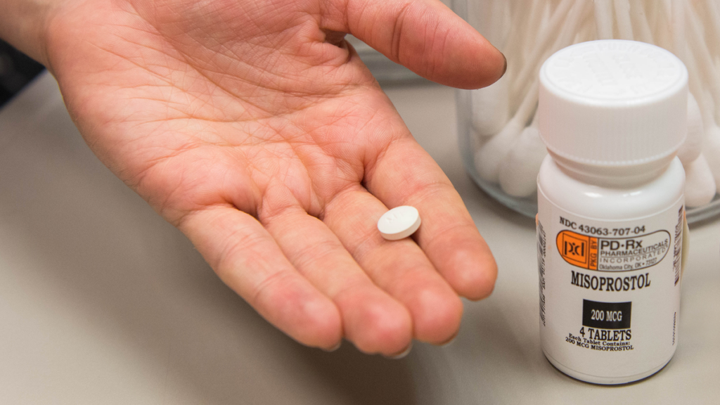 A hand holds out a white pill next to a white prescription medication container.