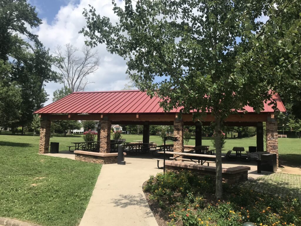 A picnic shelter at Ritter Park in Huntington. It's a sunny, cloudy day with green leaves on the trees. The picnic building has a red roof.