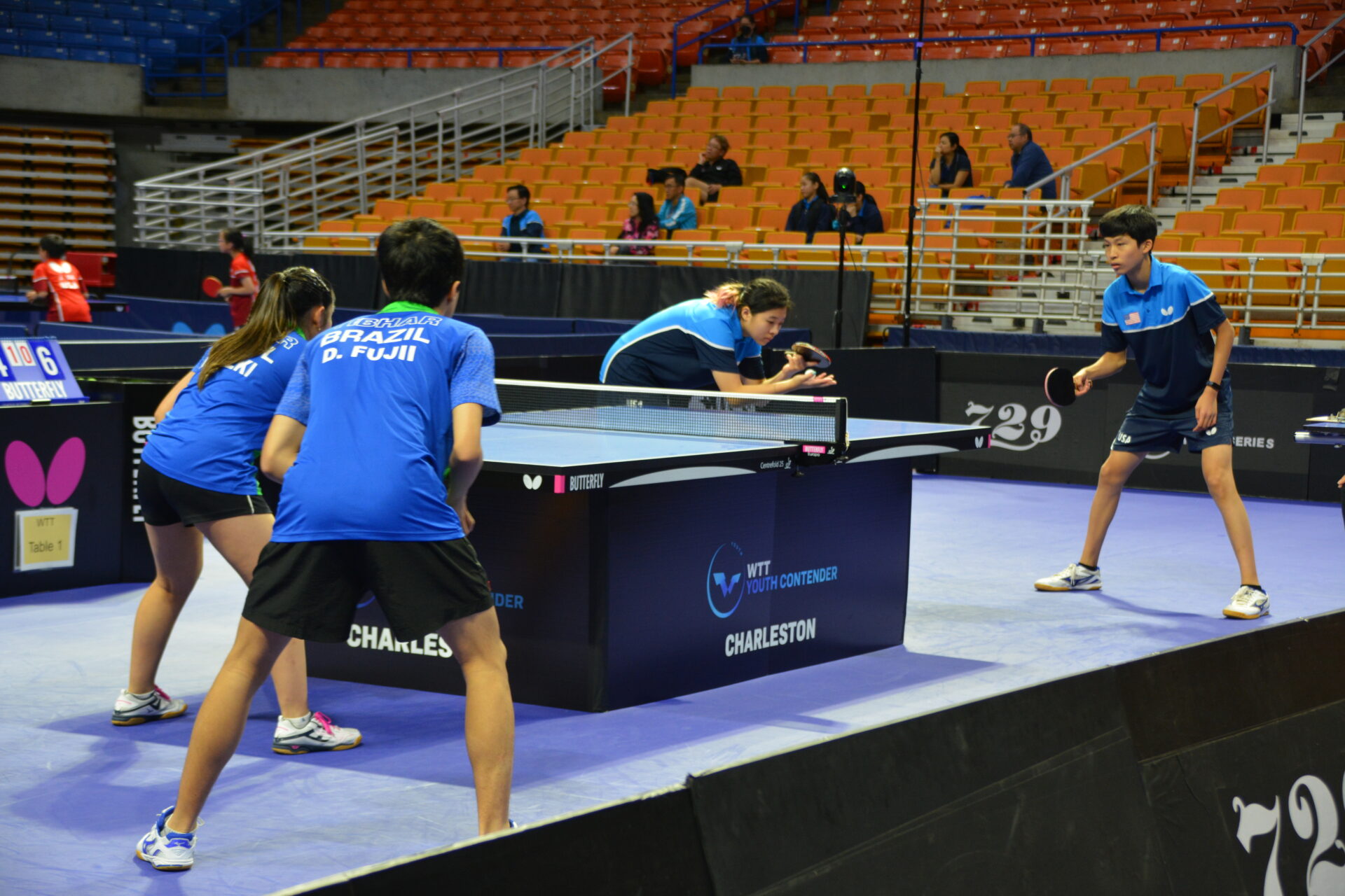 four young table tennis players face off in table tennis individual's world championship. They are all four in blue and black uniforms, playing mixed doubles. One boy and girl on each side of the table. On the far end of the table the girl is about to serve the ball. Other players are in wide stance ready to start play.