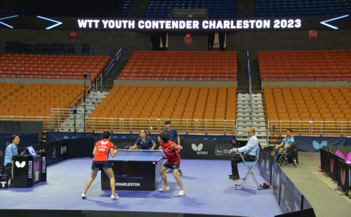Four players face in mixed doubles Table tennis mathc. The first doubles team is in red and is about to recieve the ball that the other team in blue just served over to them. The boy on the red doubles team has his paddle flexed to hit the ball that is up in the air.