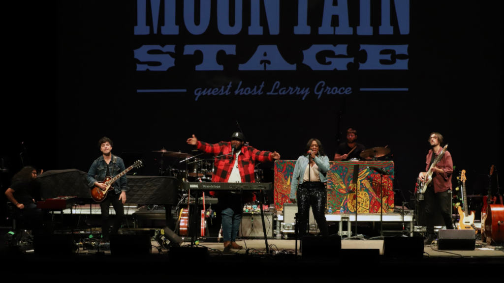 Four people perform on a stage with the words "Mountain Stage" in large letters behind them.