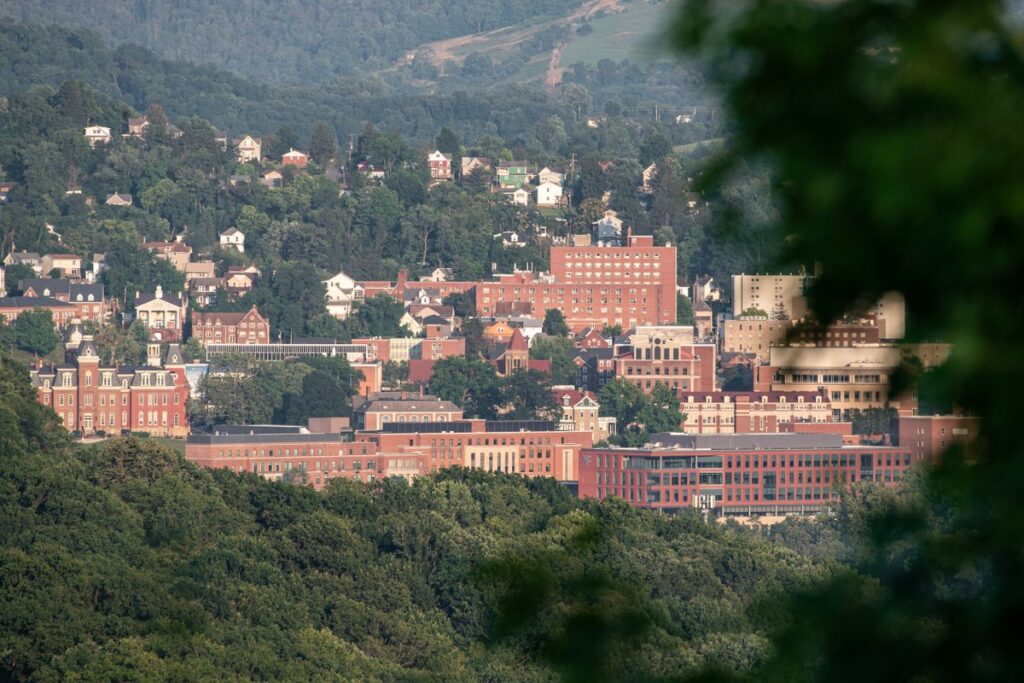 A long-distance image shows downtown Morgantown through the leaves of a tree. Nestled within verdant green, rolling hills are several red-brick buildings.