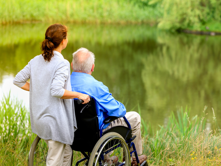 A man and woman look at a pond. The woman is pushing the elderly man's wheelchair.