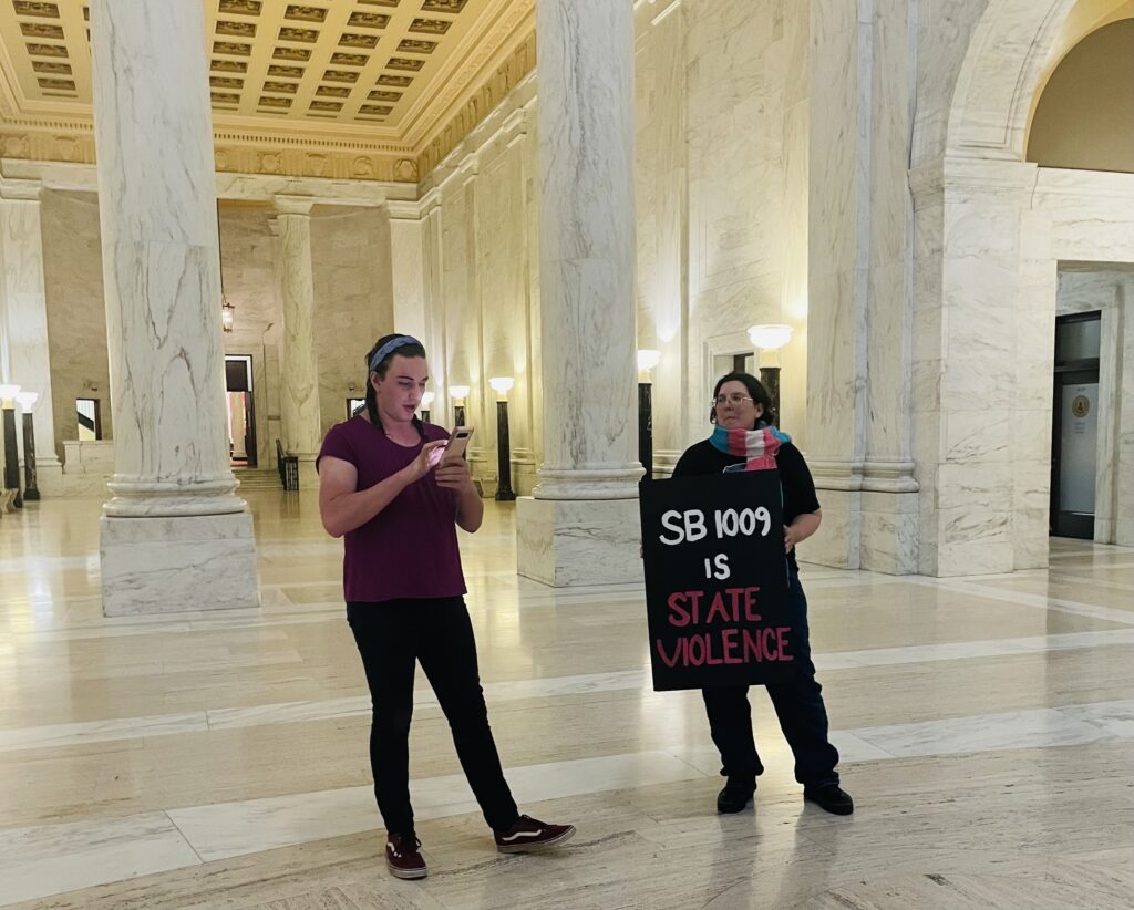 Two trans activist stand in the capitol building. Two white imposing columns stand behind them. One of the activists is holding a sign saying, "SB 1009 is State Violence." The woman next to them is reading from a phone.