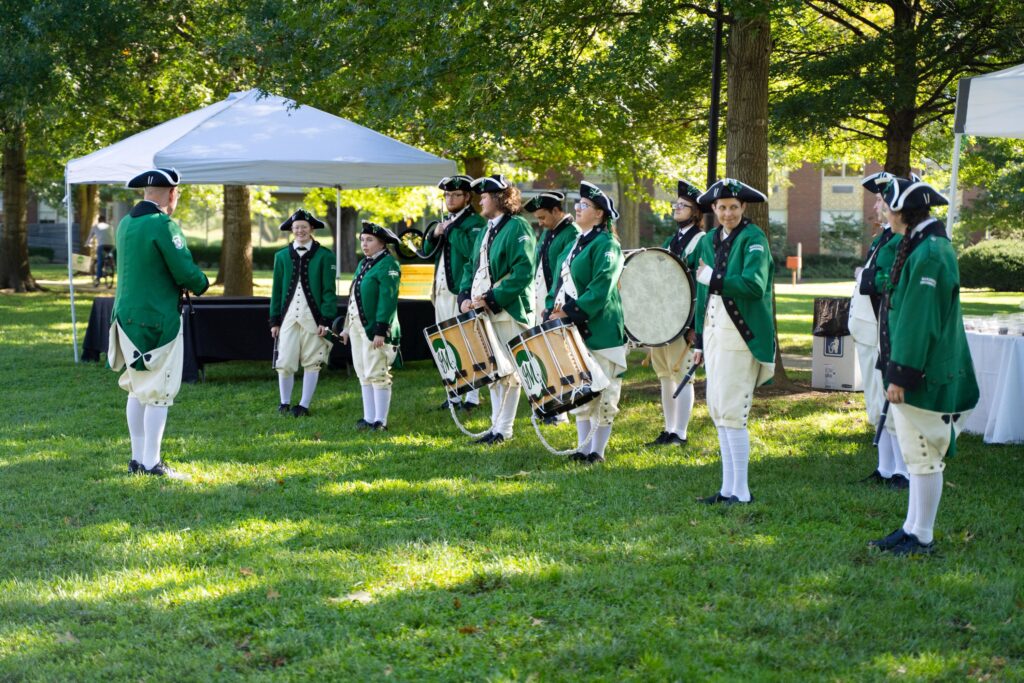 A group stands holding drums and flutes in 18th century green and white uniforms.