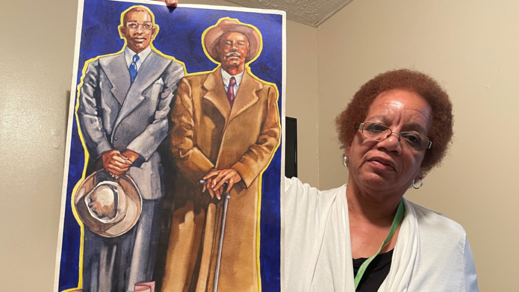An older Black woman wearing glasses holds up a painting of two Black men dressed in formal coats. One man holds a cane and wears a hat, while the other man holds his hat in his hands.