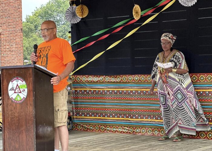 A man speaks at a podium. Behind him stands a woman dressed in traditional African dress. There are streamers hung and other decorations.