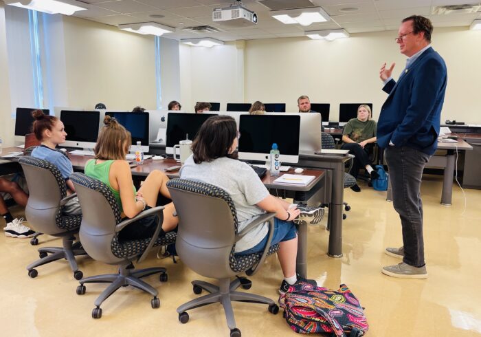 Trey Kay is seen speaking to a classroom of college students. Kay is standing, while the students all sit at computers in a computer lab.