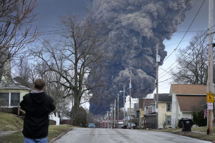 A dark plume of smoke is seen in the distance. A man, with his back to the camera and wearing a black hoodie, looks on as the smoke plume rises high in the sky.
