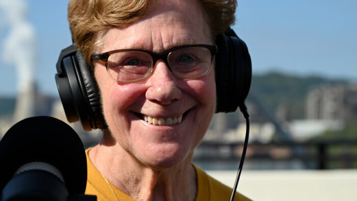 A smiling woman is shown wearing over the ear headphones, glasses, and is holding a microphone. She wears a yellow shirt.