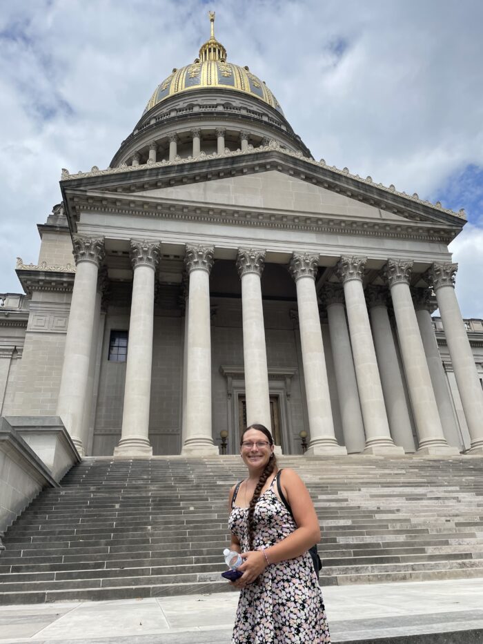 A woman in a dress stands on the steps of the West Virginia State Capitol. The sky looks overcast.