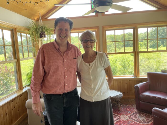 Trey Kay stands with a woman wearing a white blouse, glasses and black skirt. They stand in a large sun room with several windows and comfortable looking chairs.
