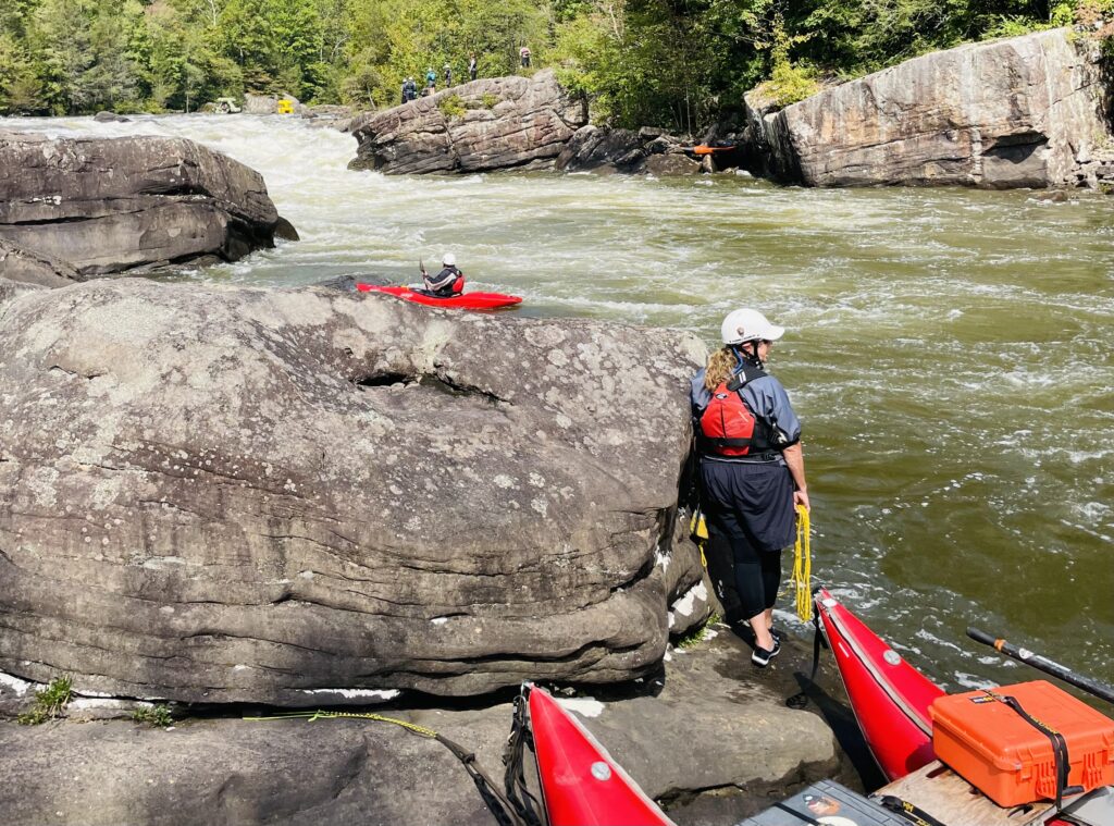 Upstream view of a rapid on a river. There are giant rocks along the river and a woman wearing a life jacket facing away from the camera holding rope.