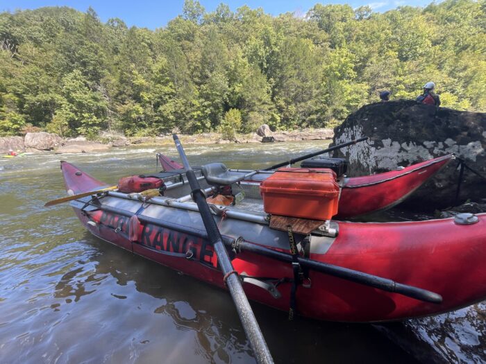 Red Cataraft is parked along the side of the river. It reads National Park Service. It has a mounted oar frame.