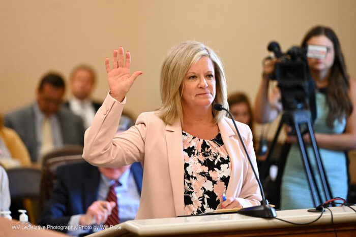 A woman in a light pink blazer over a black blouse with a pink and white floral pattern holds up her right hand while standing at a lectern with a microphone. A crowd, including a television camera, can be seen behind the woman.
