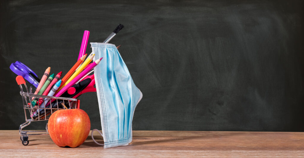 School supplies are seen on a desk in front of a chalkboard as a protective face mask hangs from a pencil.