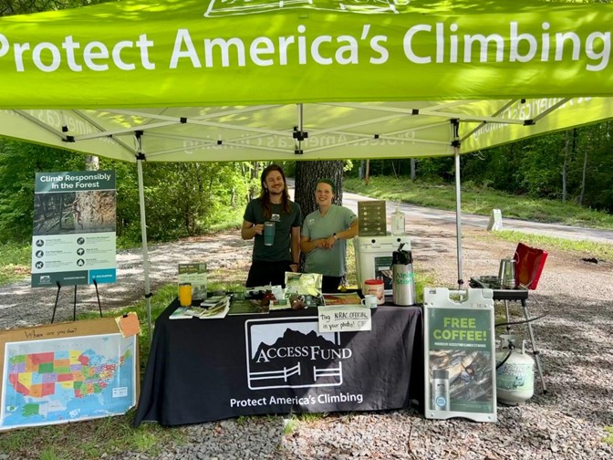 Ambassadors pose for a picture under a green pop-up tent that reads "protect Americas climbing" . The have other little signs against their table and different pamphlets and literature on their table. They also have a poster hanging with all the different counties of the state. It's a sunny day.