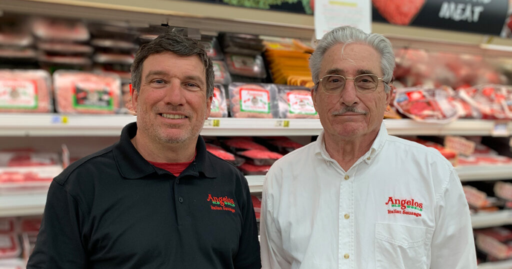 Two men stand next to each other in front of packages meat in a grocery story.