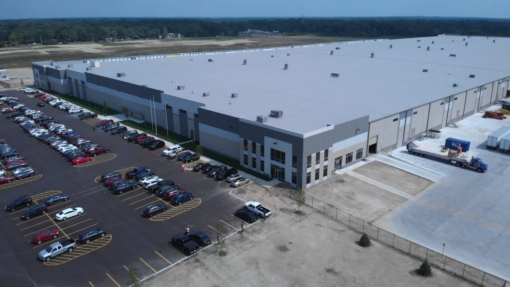 An elevated view of a sprawling distribution center with a flat roof and an employee parking lot in the front of the building.