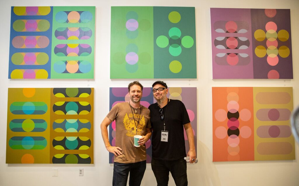 Two men, one wearing a brown Ween shirt and holding a coffee cup, the other wearing a black shirt, glasses, and cap, stand in front of a wall of multicolored canvases with symmetrical prints on them. Both men are wearing lanyards.