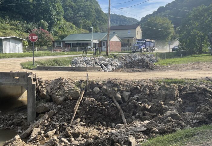 A dump truck drives along a dusty road. Closer up there is a bridge with wooden debris all around it. The bridge is in disrepair.