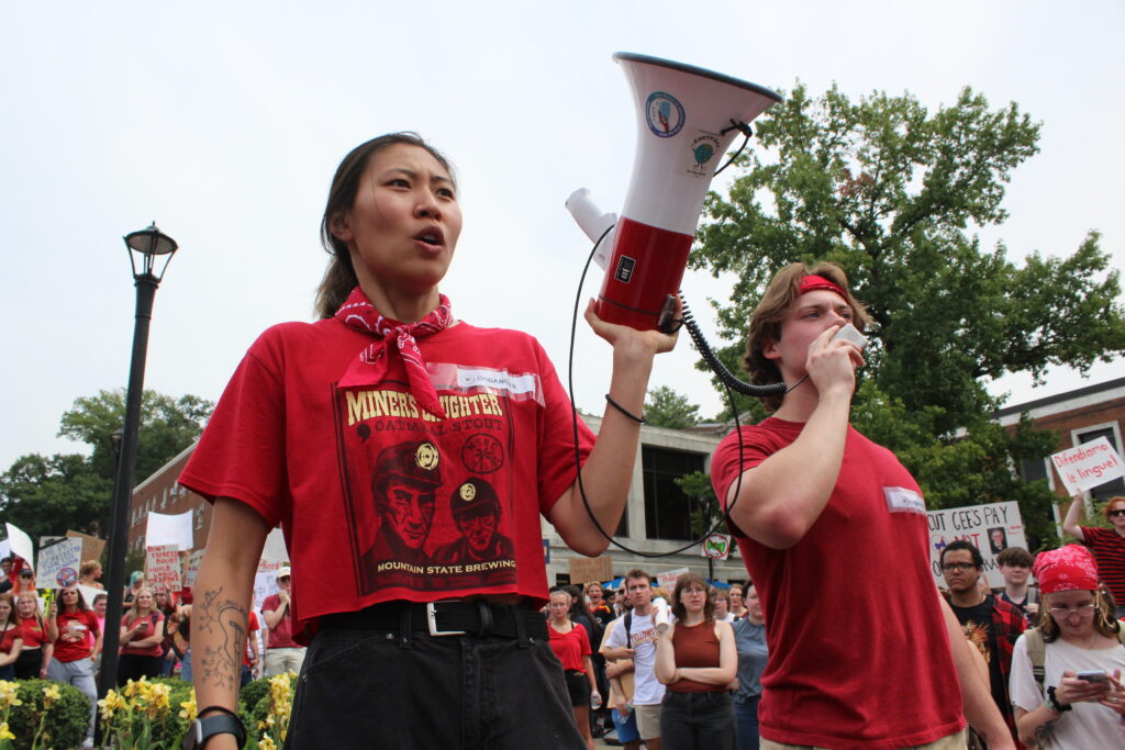 Two people stand wearing red shirts. One holds the speaker of a megaphone while the other speaks into the detached mouthpiece of the same megaphone. Behind them are arrayed more individuals, many also wearing red.