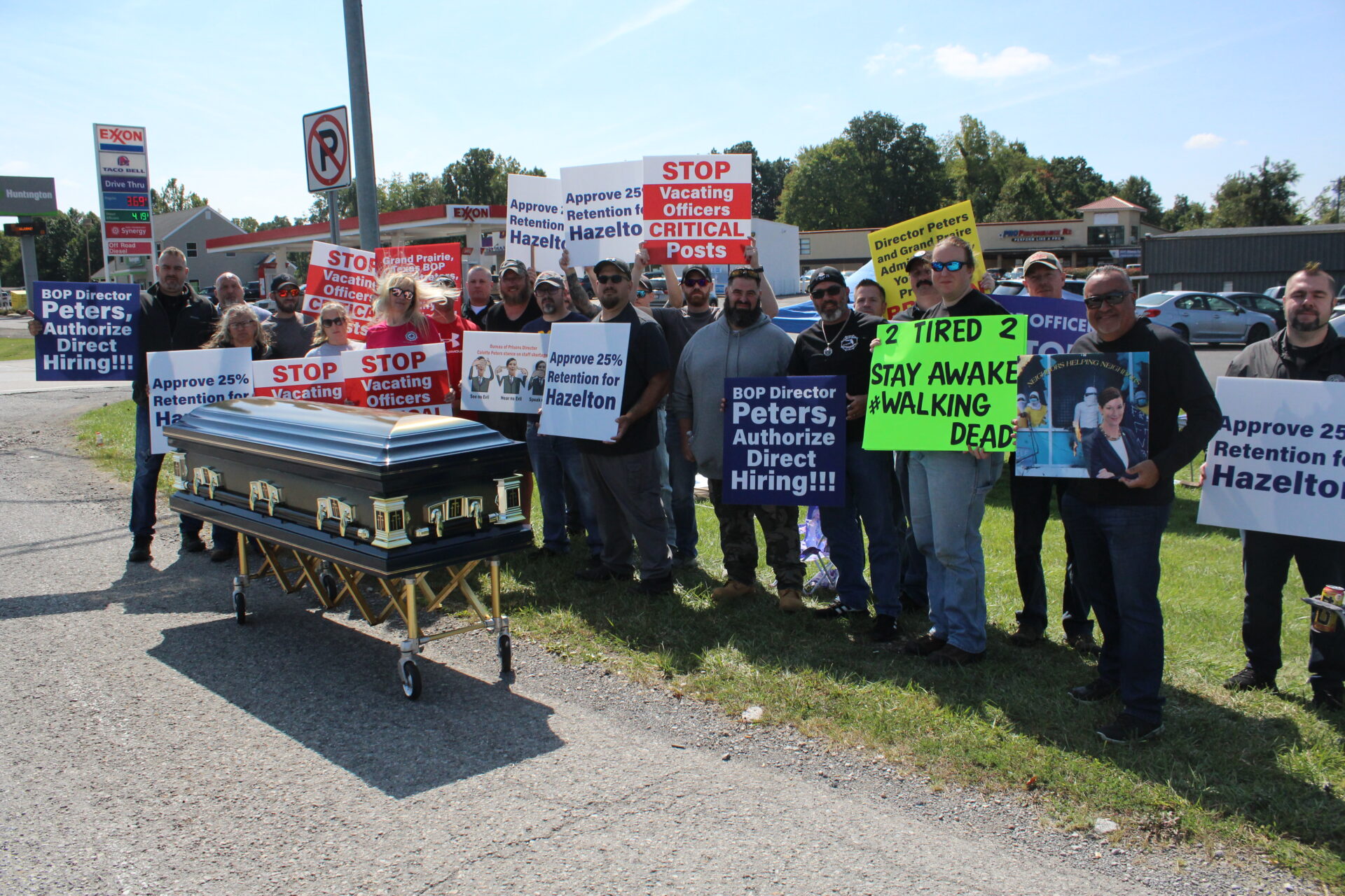 A group of people stand on the side of the road behind a casket. Many hold signs, some of which read "BOP Director Peters, Authorize Direct Hiring!!!" and "STOP Vacating Officers CRITICAL Posts" and "2 Tired 2 Stay Awake #WalkingDead"