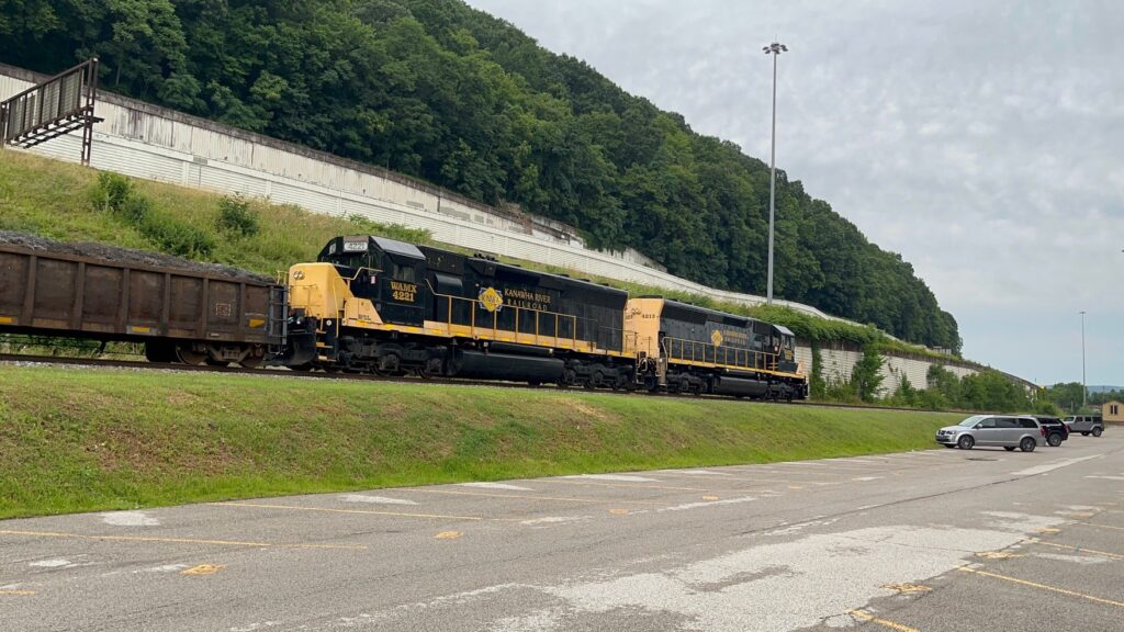 A train with two locomotives passes a parking lot with a highway in the background.
