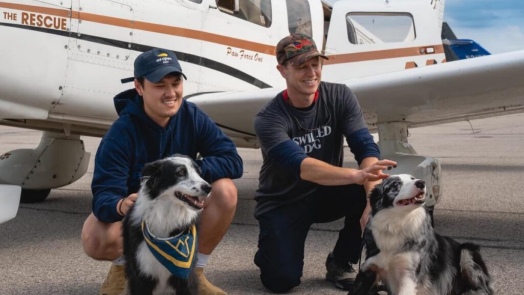 Two men kneel in front of a plane with two border collies mix breed dogs