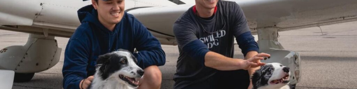 Two men kneel in front of a plane with two border collies mix breed dogs