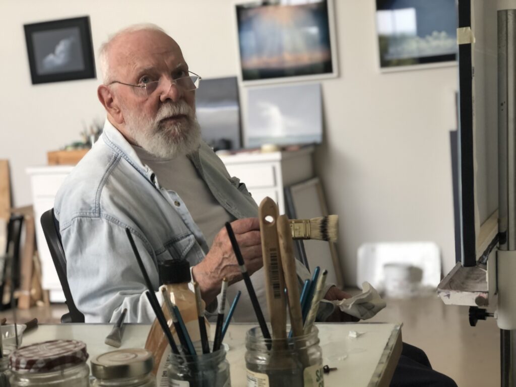Man with white hair and glasses sits at an artist desk. He is looking toward the camera. There are paint brushes on the foreground of the photo, and paintings in the background.